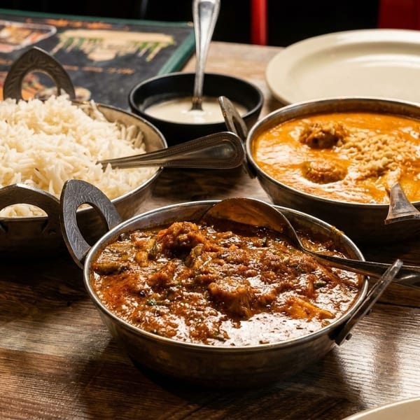 Three bowls of Indian curry and rice, with a side of raita, on a wooden table.