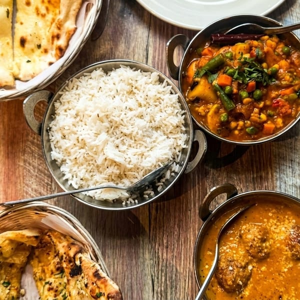 Overhead view of Indian dishes: naan, rice, vegetable curry, and kofta curry on a wooden table.