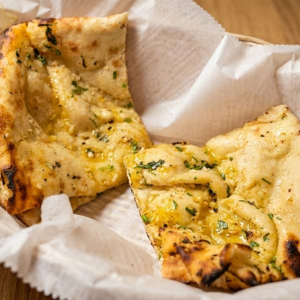 Two pieces of golden-brown garlic naan bread with herbs in a white paper-lined basket.